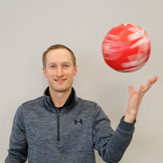 Jeff smiling at camera and throwing a red SOMN soccer ball in the air. Wearing a gray quarter zip sweatshirt