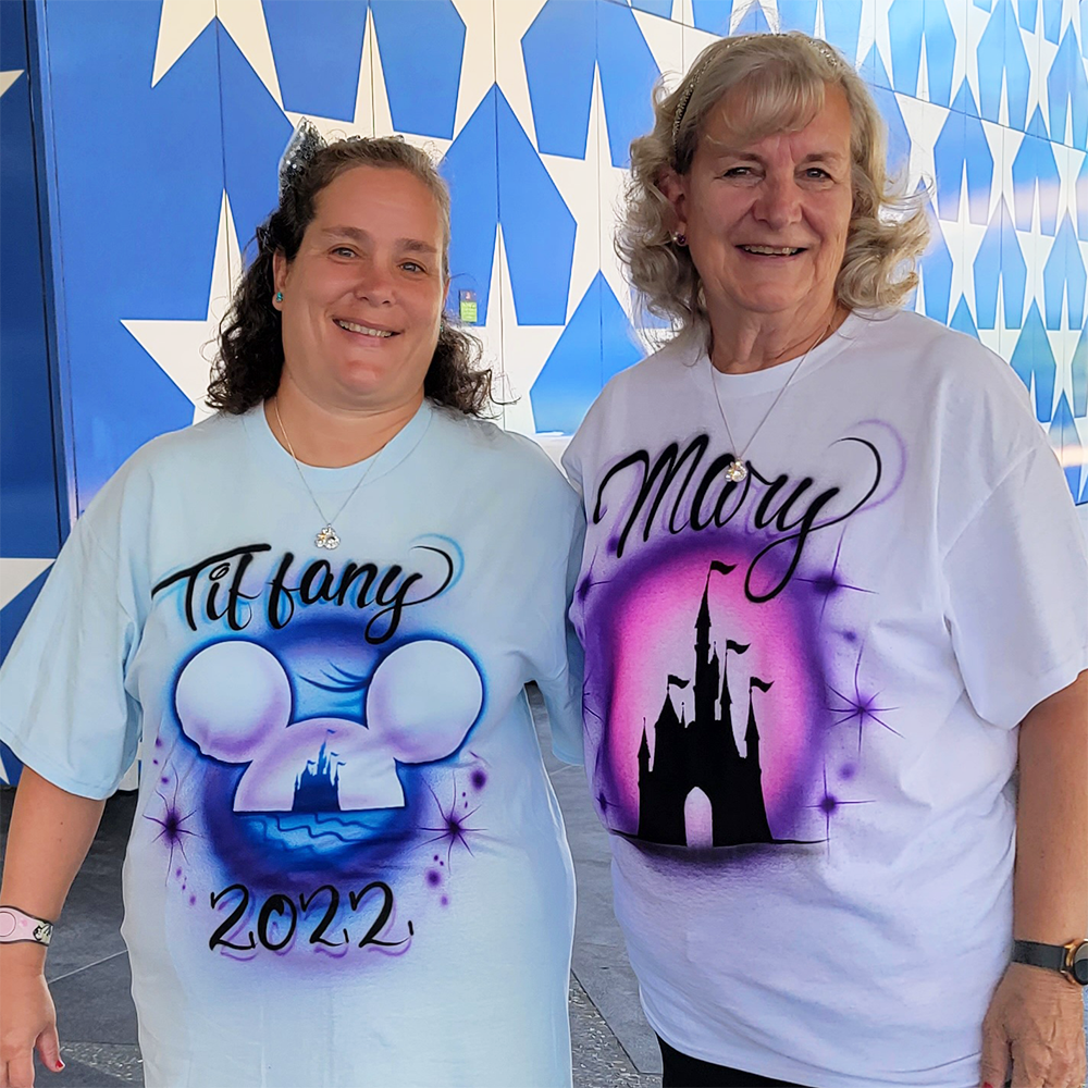 Tiffany and her mother Mary, both wearing T-shirts featuring Disney imagery and their names, pose and smile in front of a blue wall covered with white stars