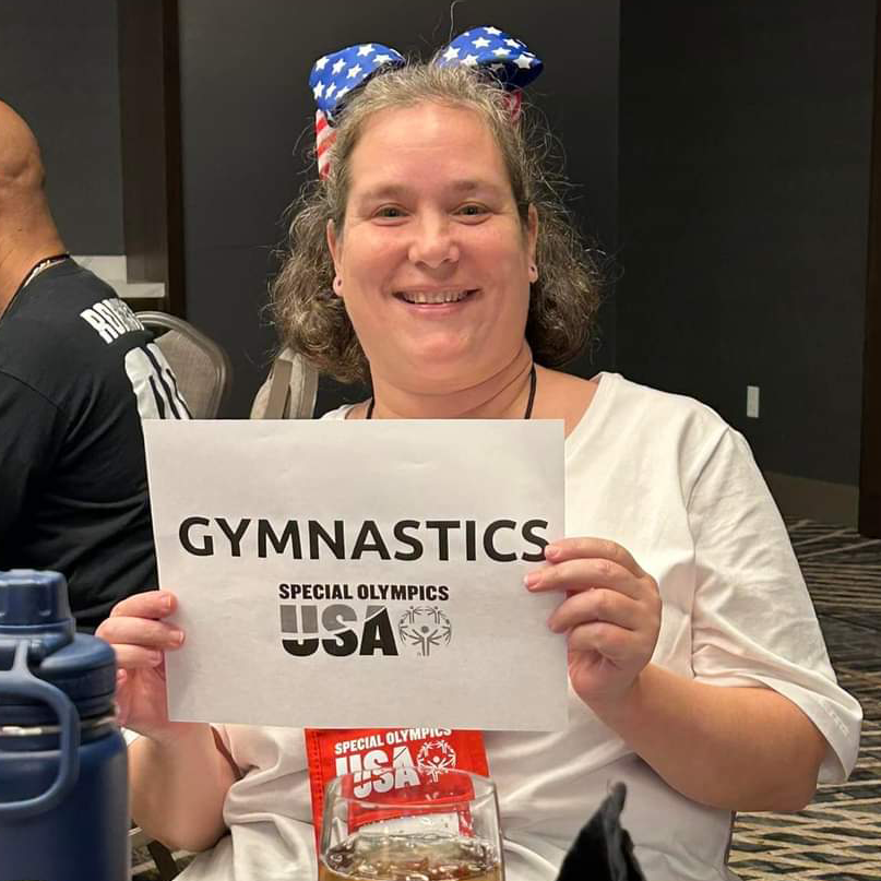 Tiffany, wearing a big bow in her hair printed with the American flag, smiles at the camera and holds up a paper sign that says Gymnastics Special Olympics USA